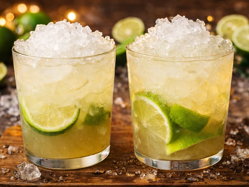Dois copos de caipirinha com limão e gelo triturado sobre uma bancada de madeira